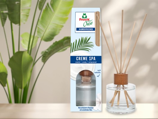 A room fragrance glass bottle with reed diffusers stands next to a Frosch Oase Orange scent sachet