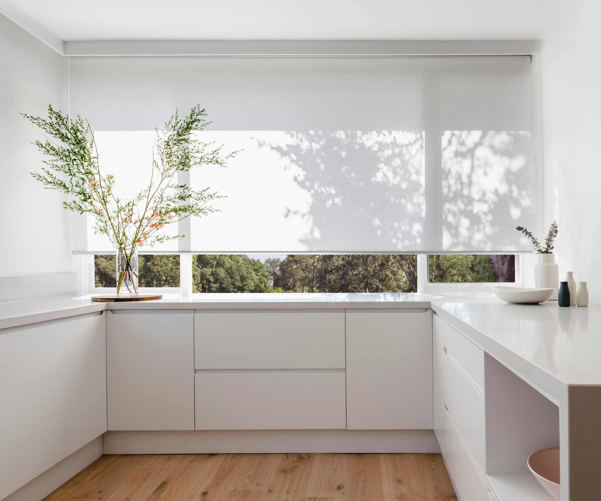 Helle Küche mit weißen Fronten vor großem Fenster mit halb geschlossenem Sonnenschutz