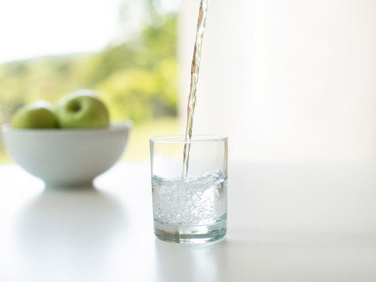 Water is poured into a glass with a bowl of apples in the ba
