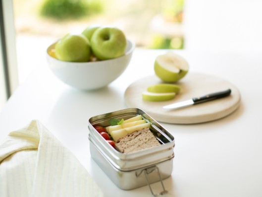 A lunch box, a bowl with apples and a cutting board with a k