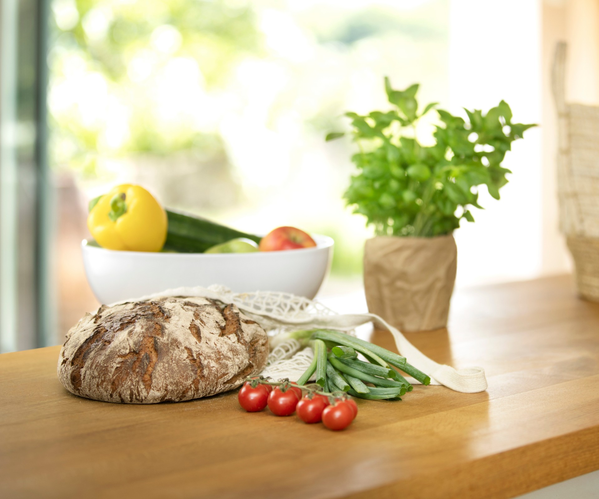 Auf einem Holztisch liegen ein Brot, Tomaten, Basilikum und eine Schale mit Gemüse
